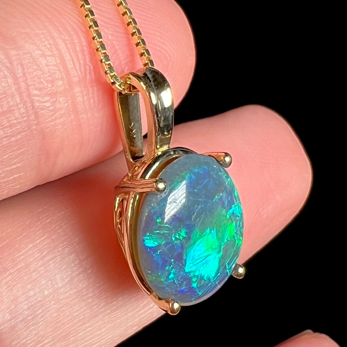 Angled close-up of 14k yellow gold Lightning Ridge black opal pendant resting on fingers, teal and green colored flashes visible in the opal