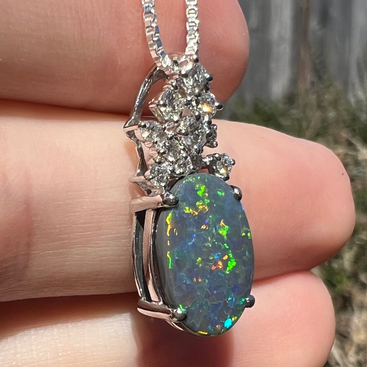 Side close-up of a platinum black opal and diamond pineapple pendant, held in hand beneath natural sunlight.