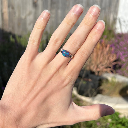 A black opal platinum ring with different shades of blue fire worn on a hand.  The opal is set East-West orientation.