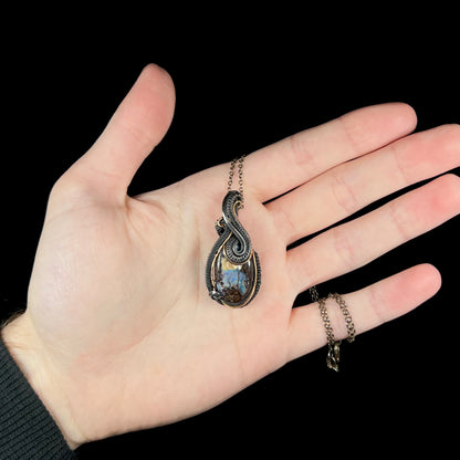 Hand holding a blackened sterling silver and gold-tone wire-wrapped necklace set with a bluish and brown Australian boulder opal.