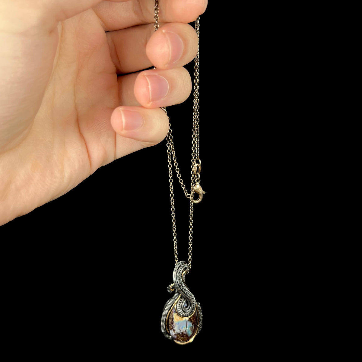 Hand dangling a blackened sterling silver wire wrapped pendant set with a boulder opal from a blackened base metal chain.