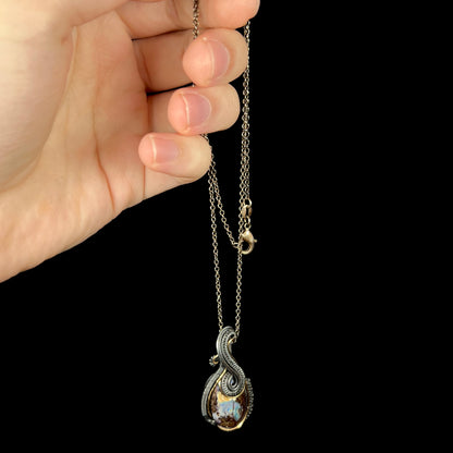 Hand dangling a blackened sterling silver wire wrapped pendant set with a boulder opal from a blackened base metal chain.