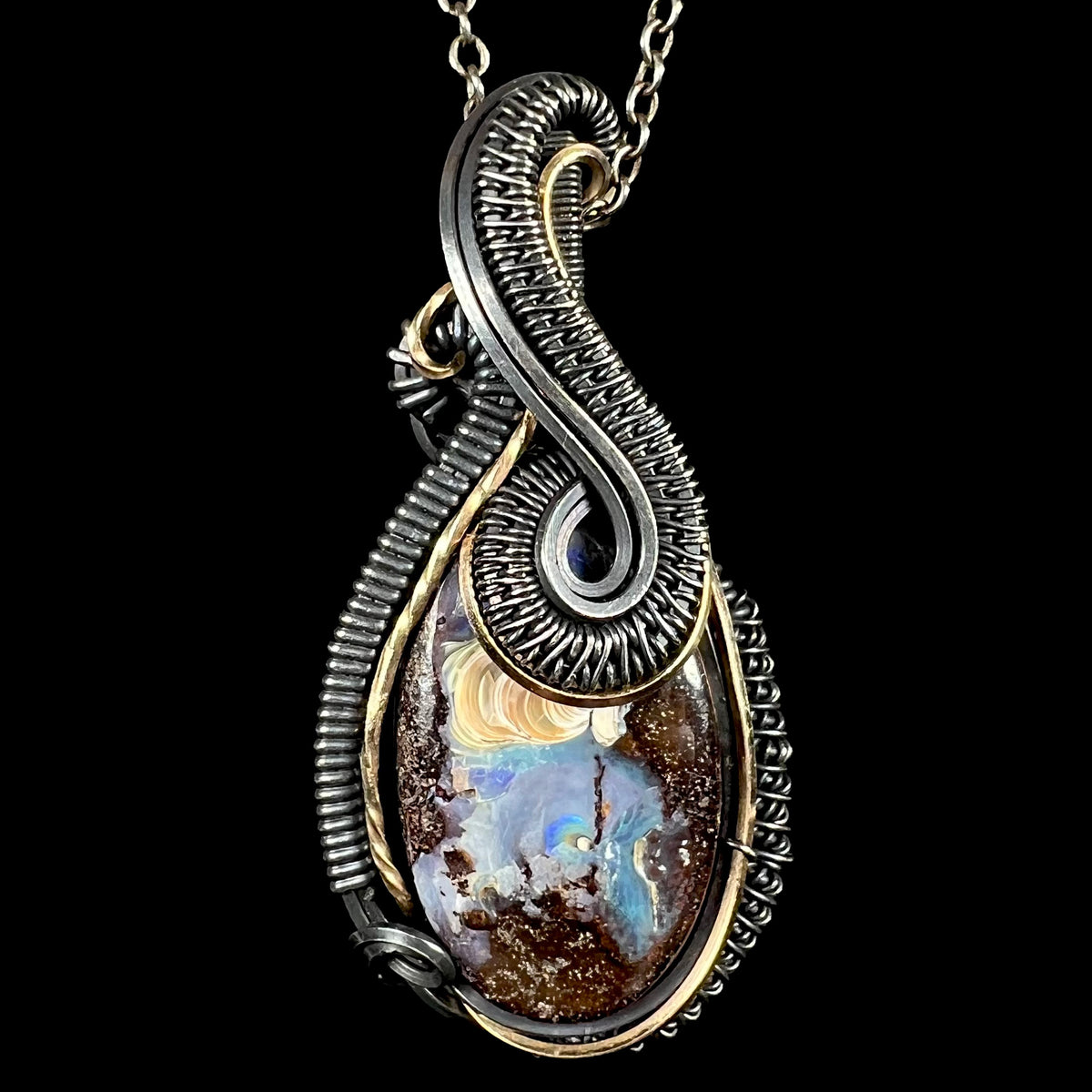 Macro close-up of teardrop-shaped pendant necklace featuring a natural boulder opal with blue, gold, and brown inclusions, set in a dual-tone blackened silver and gold wire-wrapped design, shown against a solid black background.
