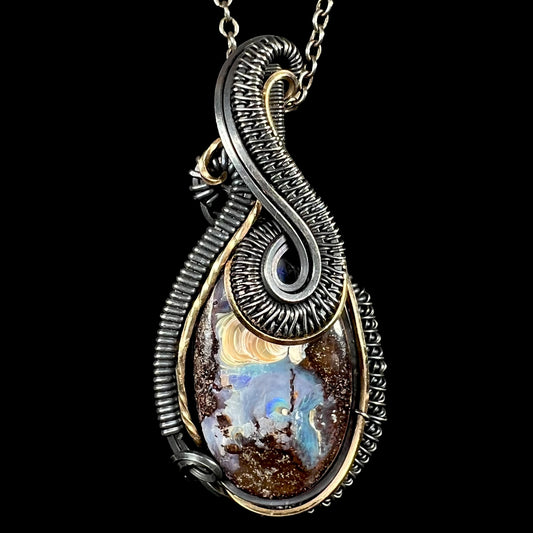 Macro close-up of teardrop-shaped pendant necklace featuring a natural boulder opal with blue, gold, and brown inclusions, set in a dual-tone blackened silver and gold wire-wrapped design, shown against a solid black background.
