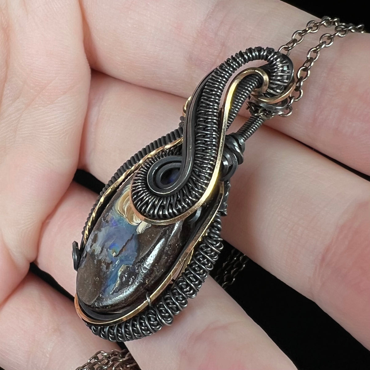 A blackened sterling silver and gold-tone wire wrapped pendant set with a natural Australian boulder opal, shown in a hand close-up from a side angle.