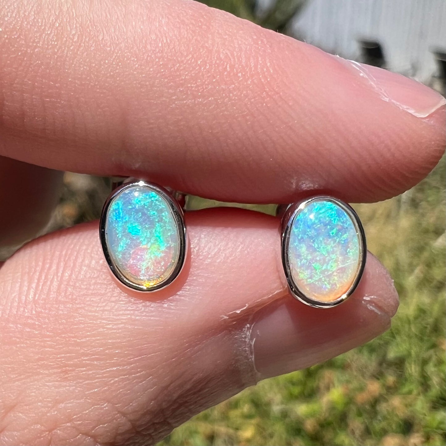 Close-up of blue Australian crystal opal stud earrings in 18k white gold, shown between two fingers in natural light.