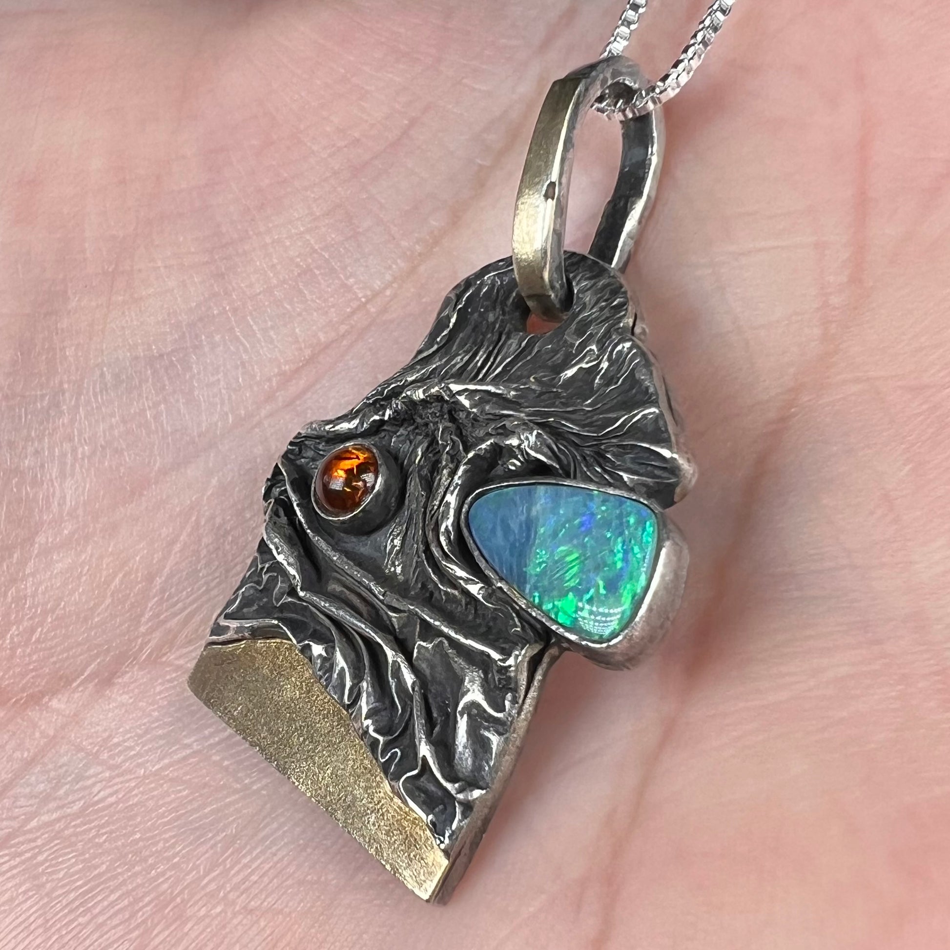 A brutalist-style blue Australian opal doublet and amber-accented men's pendant in sterling silver and 18k gold, shown close-up from an angle in a hand.