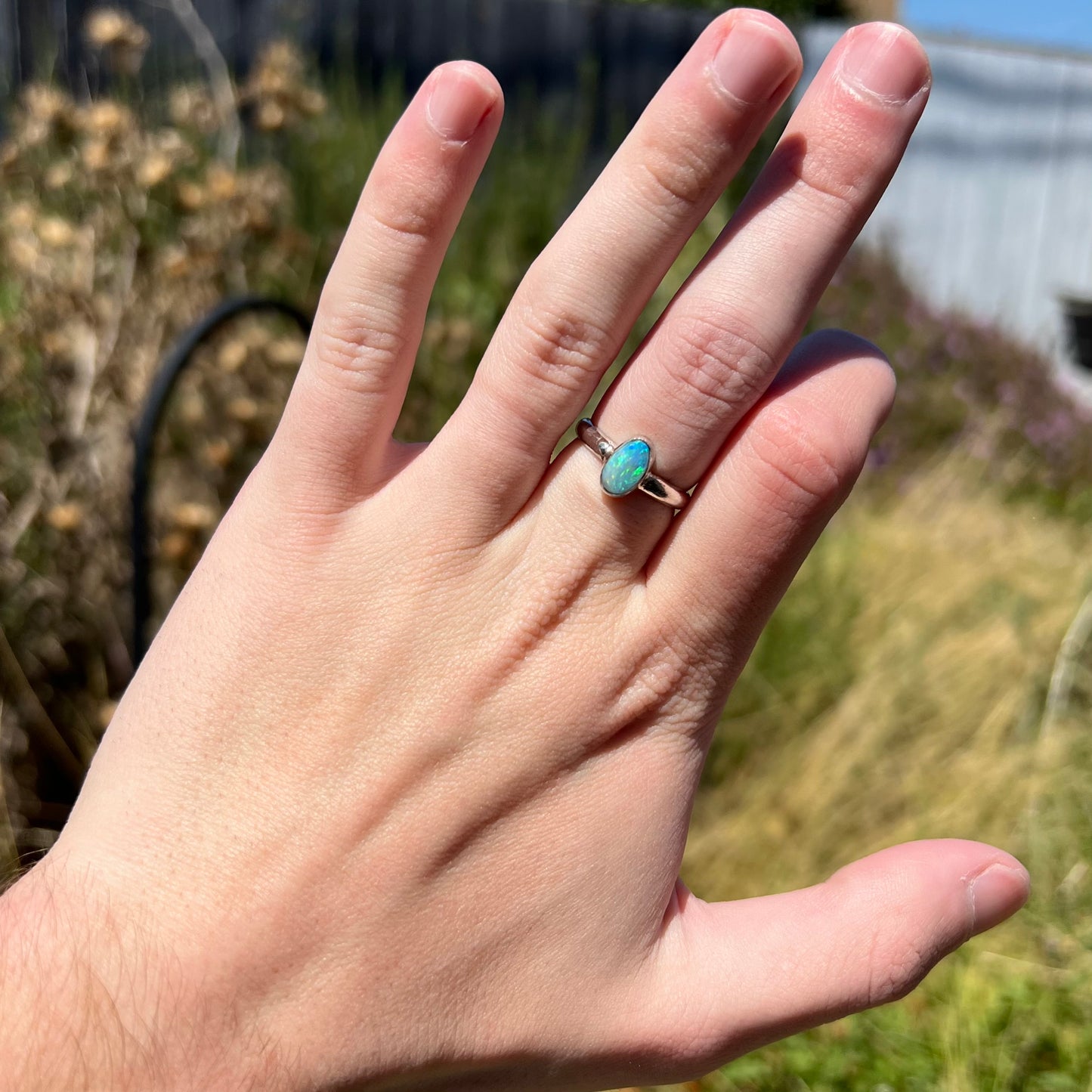 Hand wearing a blue Australian opal ring made from sterling silver in natural sunlight.  A blurred outdoor backdrop is seen in the background.