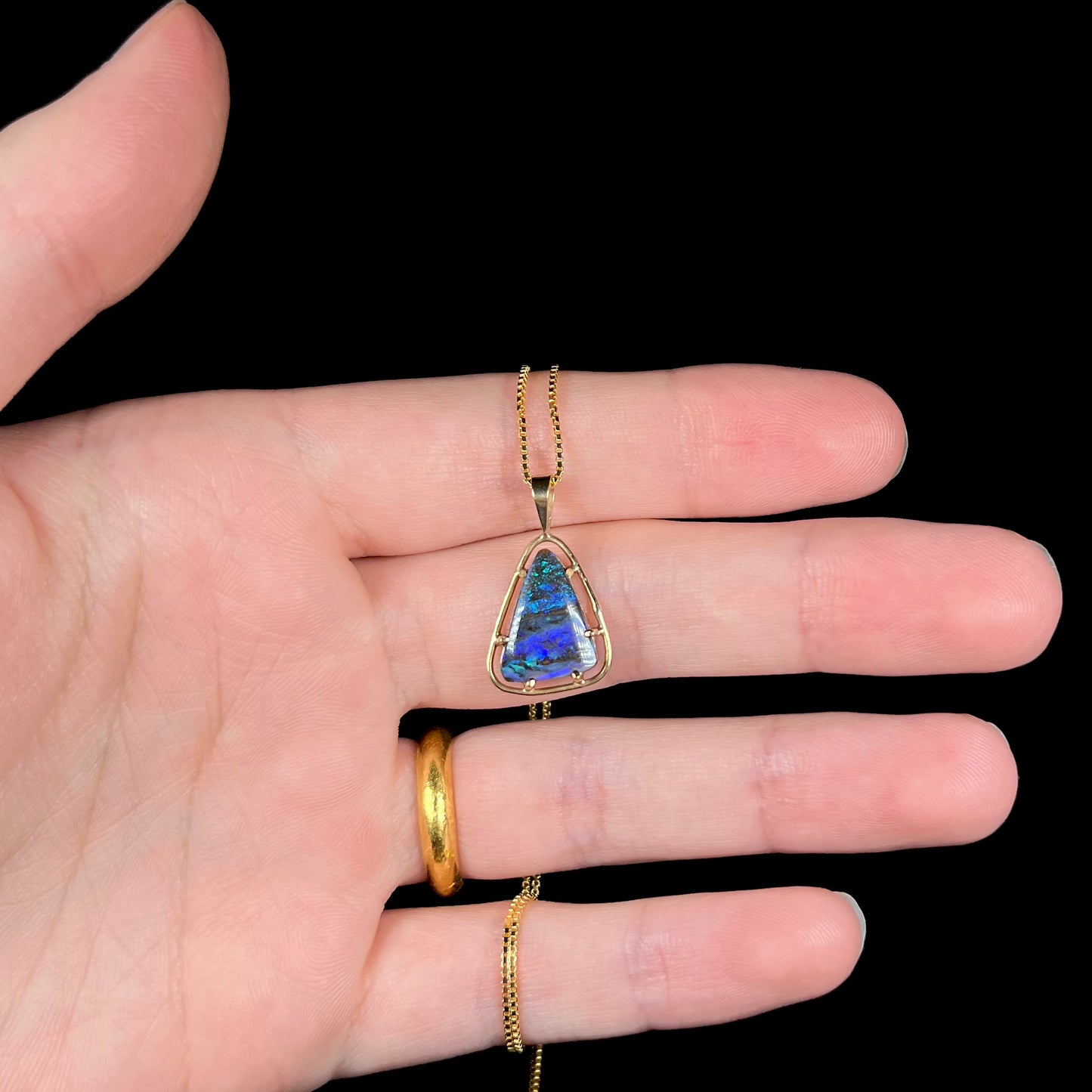 A dainty, blue Duck Creek opalized wood necklace in 9k gold, shown in a hand against a black background.