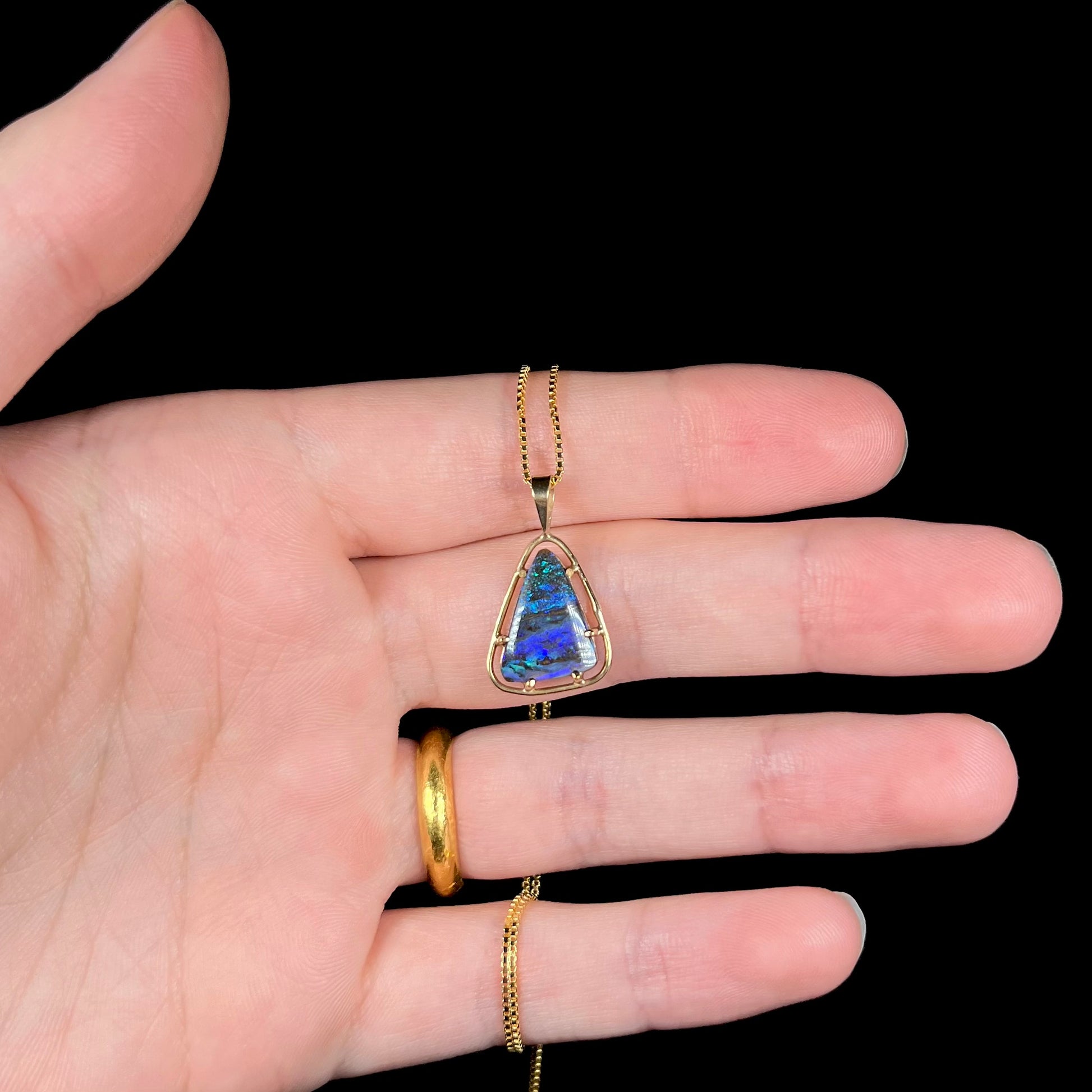 A dainty, blue Duck Creek opalized wood necklace in 9k gold, shown in a hand against a black background.