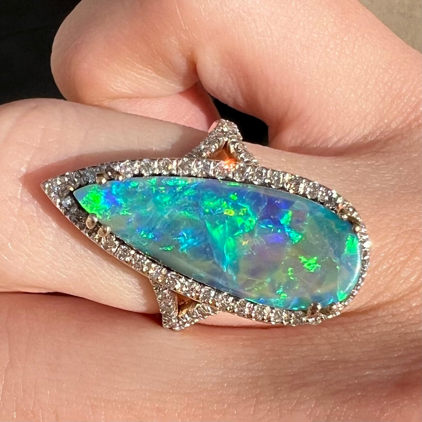 A brilliant blue boulder opal and diamond halo engagement ring by OpalPop, shown close-up on a finger in sunlight.