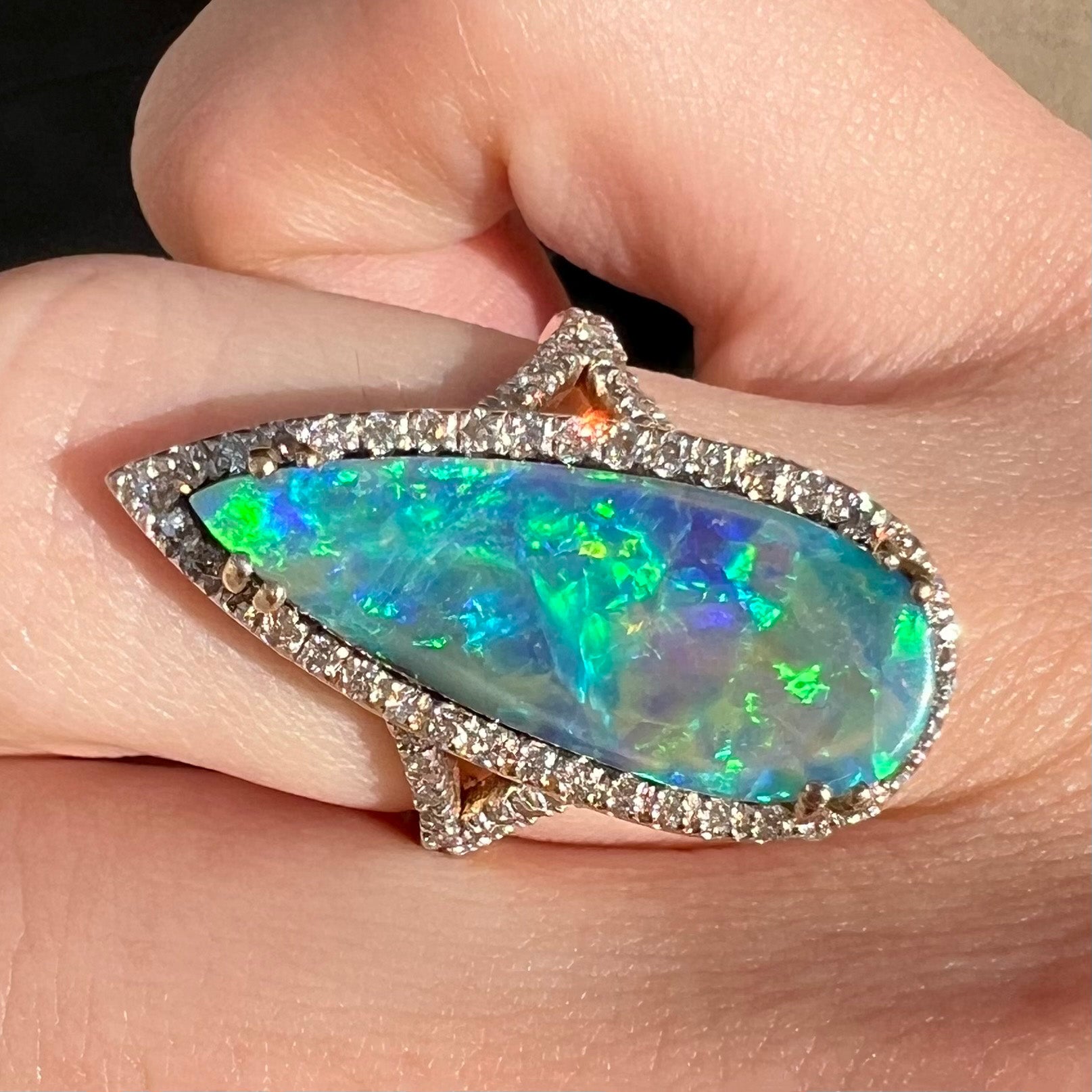 A brilliant blue boulder opal and diamond halo engagement ring by OpalPop, shown close-up on a finger in sunlight.