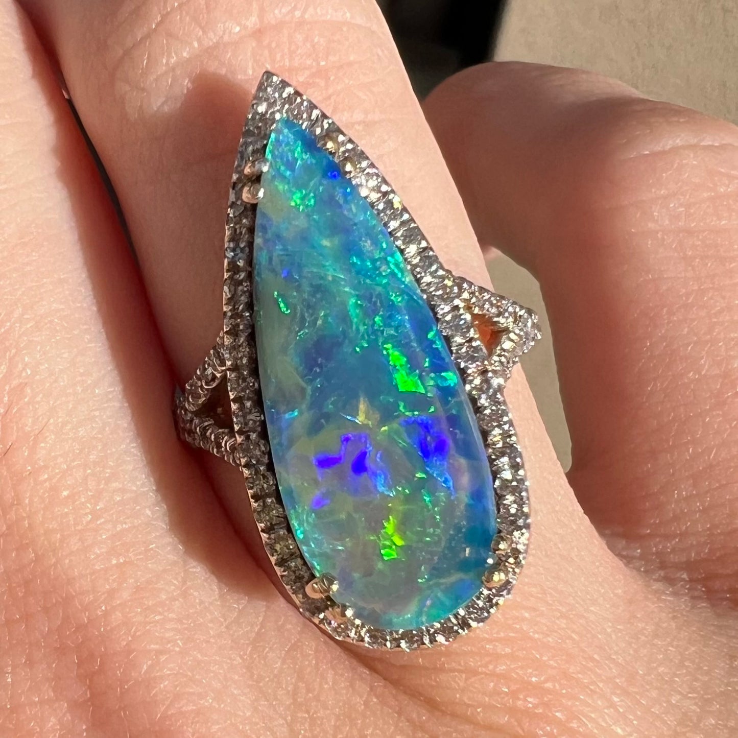 A blue boulder opal and VS2 diamond halo ring worn on a finger and shown close-up in the sunlight.