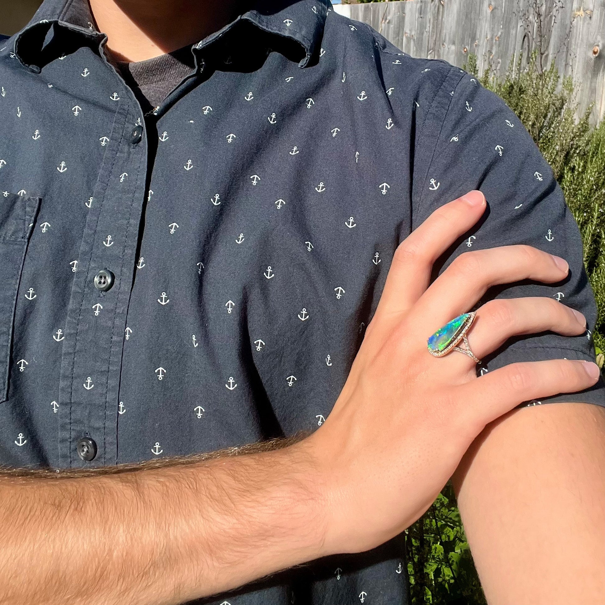 Person with blue anchor shirt resting hand on opposing shoulder while wearing a pear-shaped blue boulder opal ring in the sunlight.