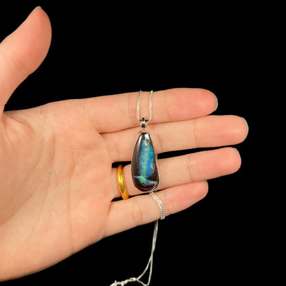 A sterling silver boulder opal necklace with blue color play, shown in a hand against a black backdrop.