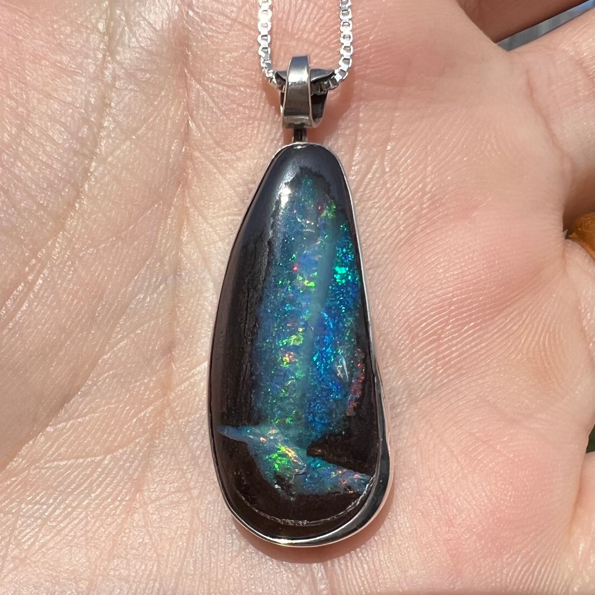 Close-up of an Australian boulder opal pendant with glittering blue and green color play, shown in a hand in sunlight.