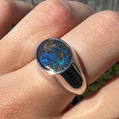 Angled close-up of a stainless steel men's Australian boulder opal ring with blue color play on a finger in sunlight.