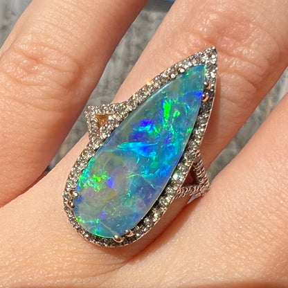 Close-up of a blue boulder opal ring with a VS2 diamond halo, worn on a finger in the sunlight.