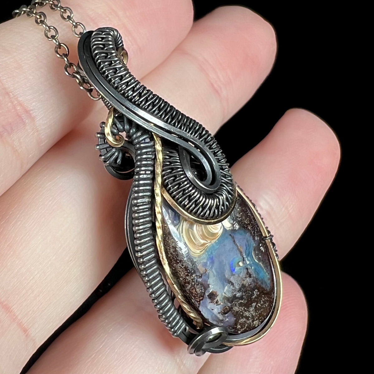 Angled close-up of a blue Australian boulder opal pendant with a two-tone blackened sterling silver and gold-tone design, held in a hand.
