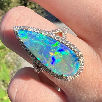 Boulder opal ring with blue flashes of color play and diamond accents, worn on a finger in sunlight and shown close-up.