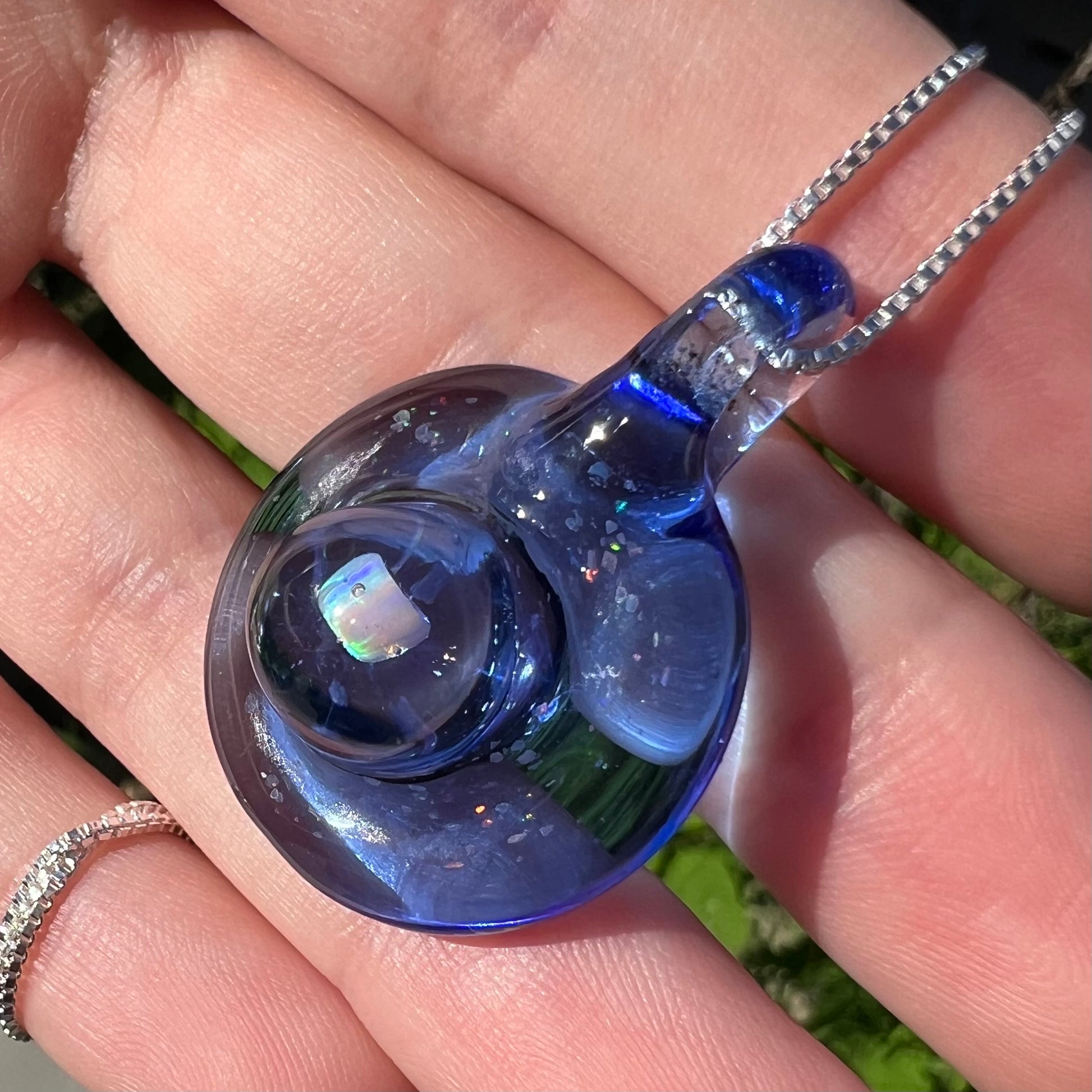 A blue glass and lab-created opal pendant shown from an angle, held in a hand in the sunlight.