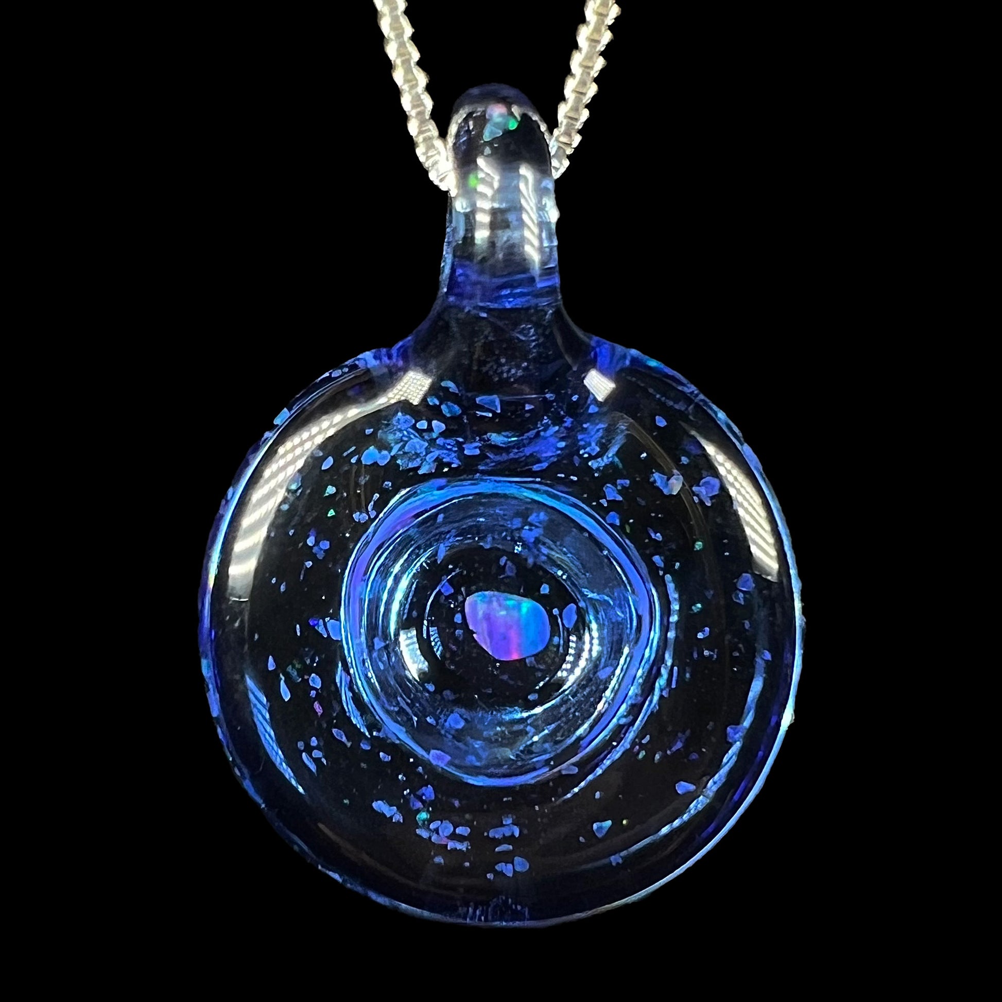 A round blue glass pendant with a rough piece of opal and opal chips floating from within, shown from the back side against a black backdrop.