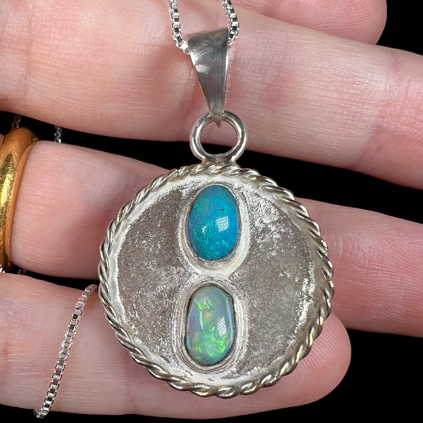Close-up of a blue and green Australian opal pendant made from textured sterling silver, shown resting on fingers.