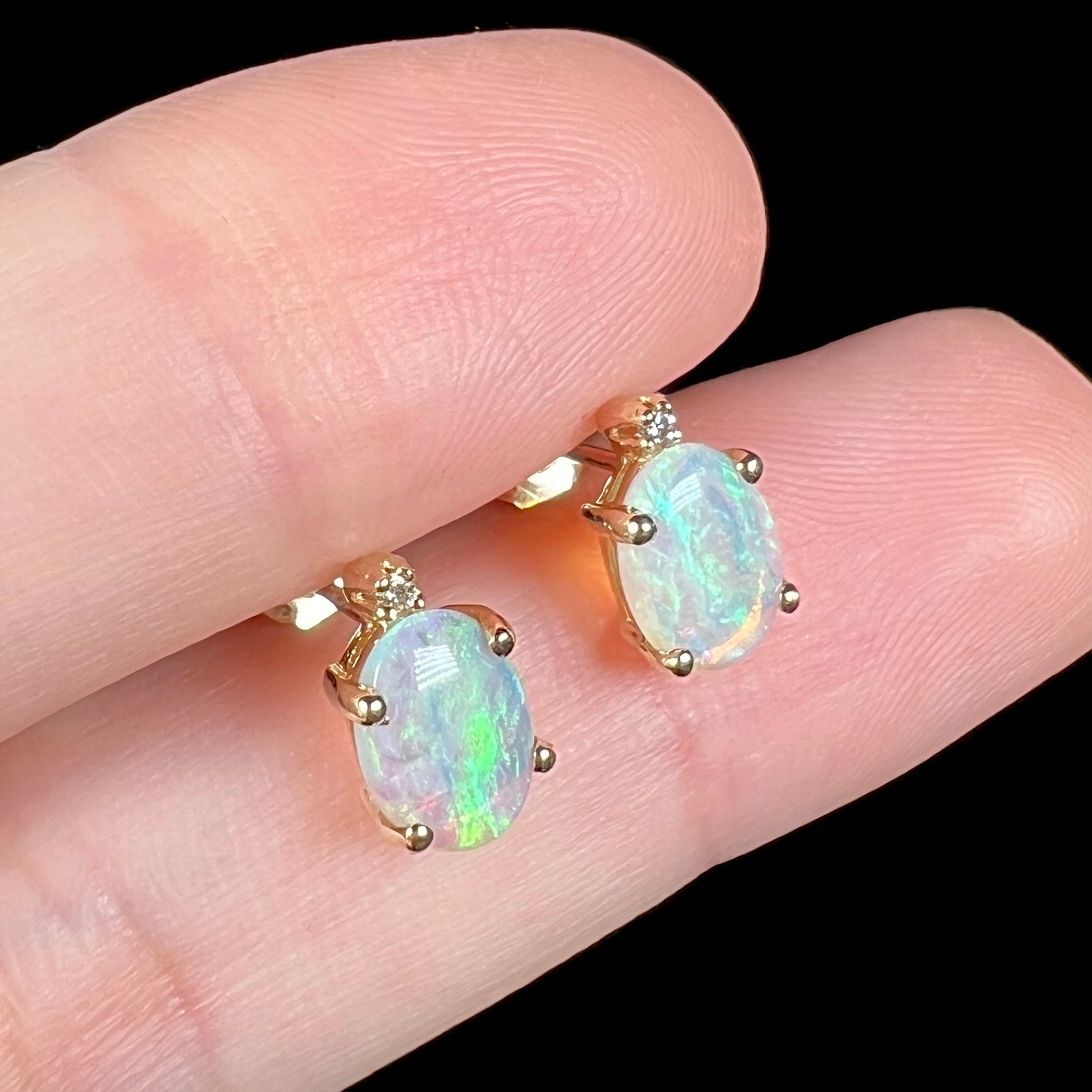 Angled close-up view of a pair of Australian crystal opal and diamond stud earrings in gold with green-blue color play shown between two fingers.