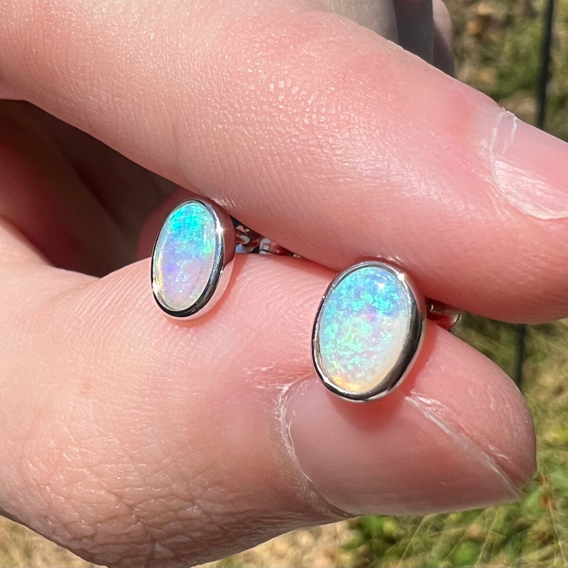 Pair of gentle blue and green Australian crystal opal earrings shown close-up in natural light, held between two fingers in the sun.