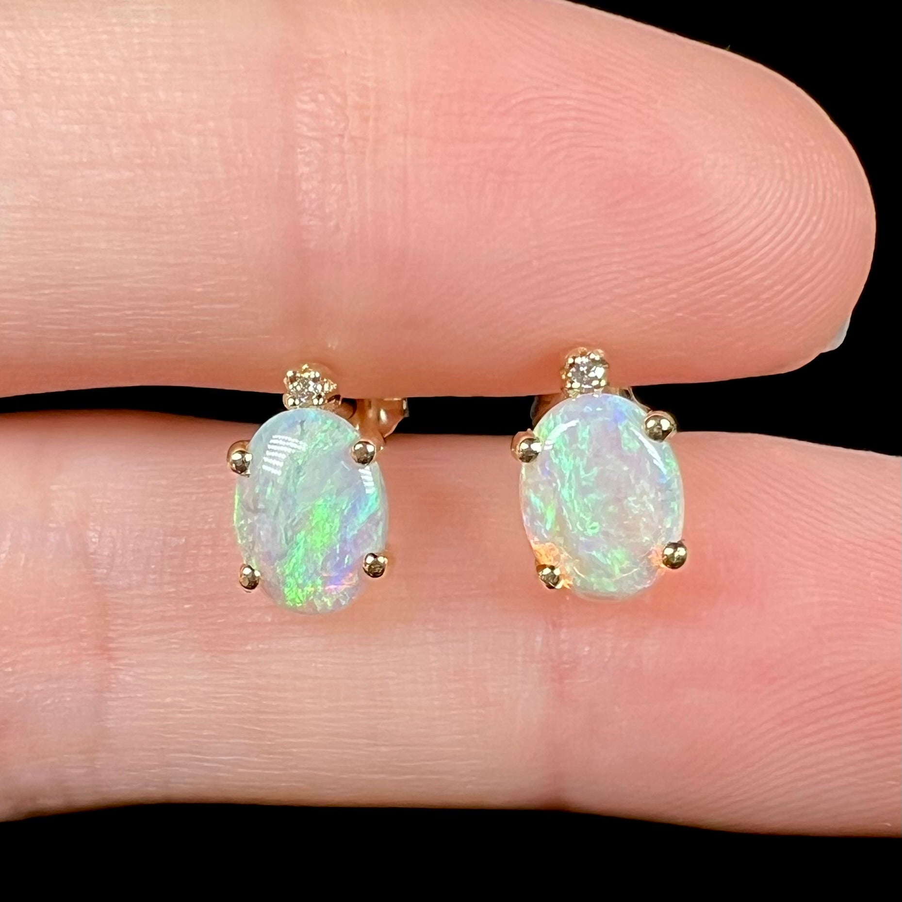 A pair of Australian crystal opal and diamond stud earrings in gold with green-blue color play shown close-up held between two fingers.