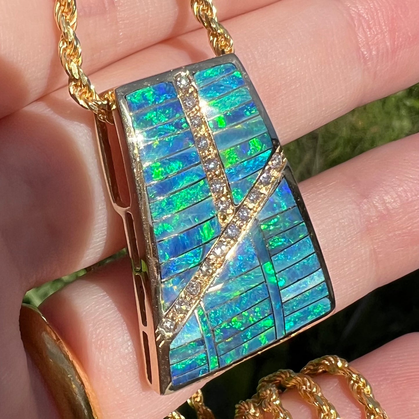 Angled close-up of a blue and green opal inlay pendant held in a hand under direct natural sunlight.