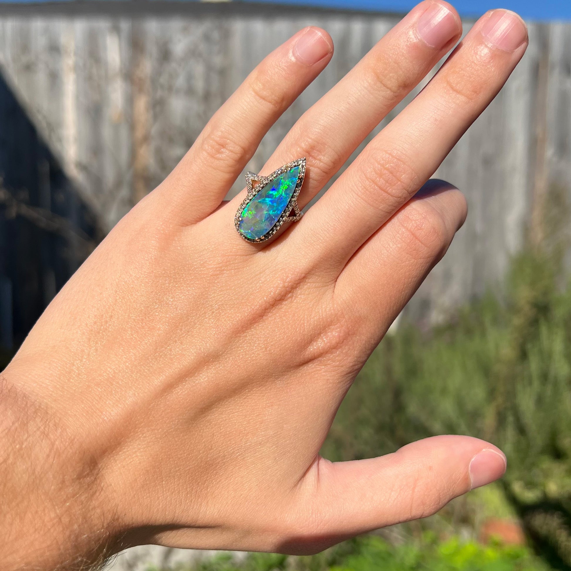 A blue-green pear-shaped boulder opal and diamond ring worn on a hand in the sunlight against a blurred outddor backdrop.