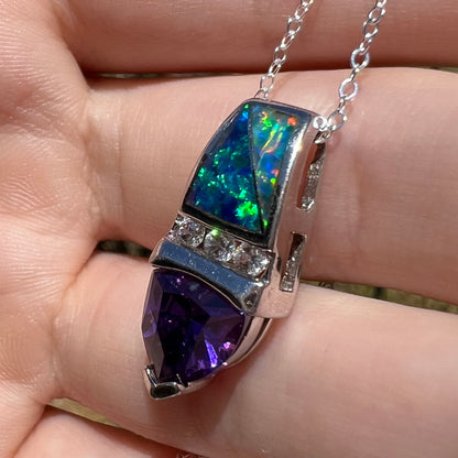 A sterling silver pendant with two pieces of blue lab-created opal inlay and a purple cubic zirconia stone drop, shown close-up from angle in sunlight against fingers.