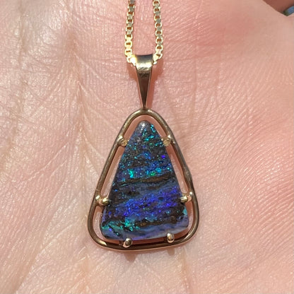 A dainty, blue Duck Creek opalized wood pendant in 9k gold, shown close-up in a hand in sunlight.