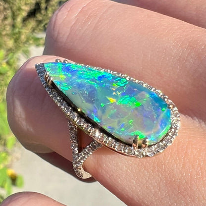 Angled sunlit close-up of a blue pear-shaped boulder opal and diamond engagement ring worn on a finger.