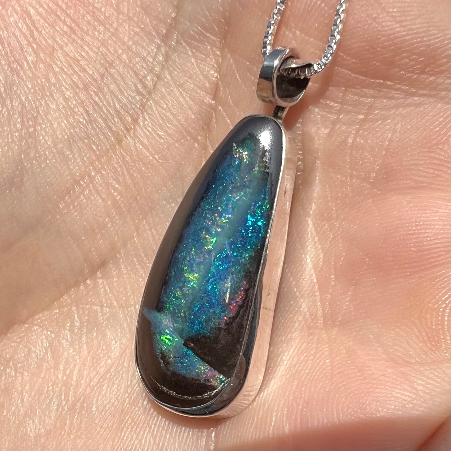 Close-up of a boulder opal pendant with striped blue and green color play, shown from an angle in sunlight.