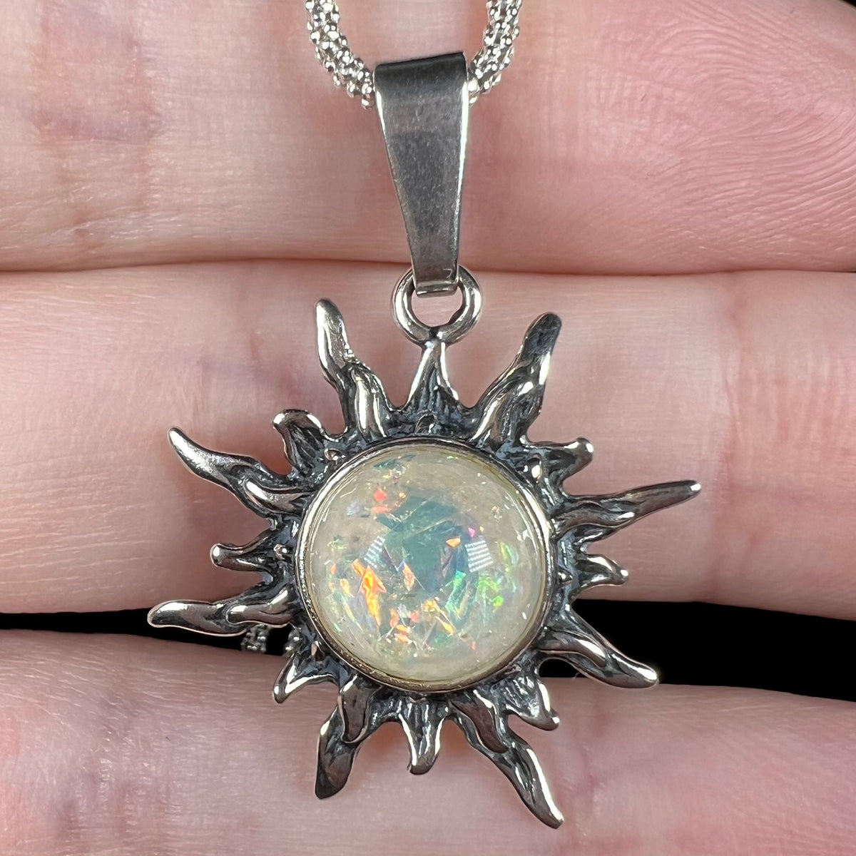 Close-up of a Boho-style sun pendant cast from sterling silver and bezel set with a round imitation opal glass stone, resting on top of fingers.