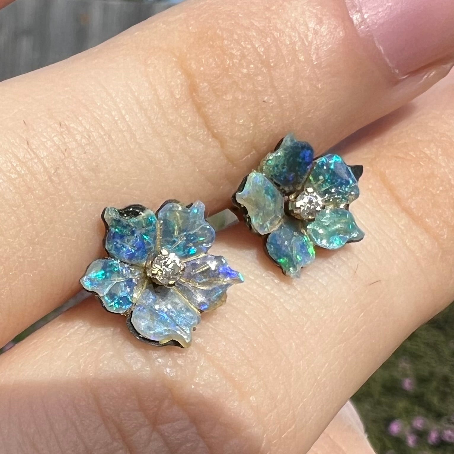 A pair of carved boulder opal doublet flower earring jackets with gold CZ studs shown from an angle in sunilght between two fingers.