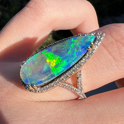 A boulder opal engagement ring with a bright green flash of color play, shown close-up from an angle in the sunlight while worn on a finger.