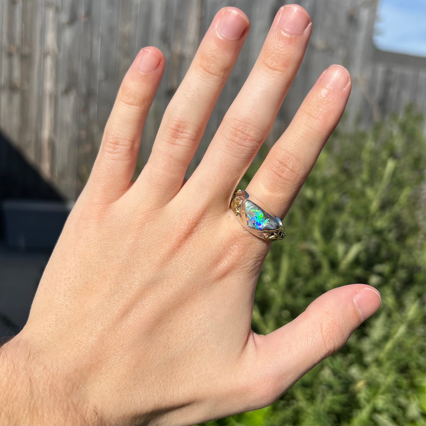 Hand wearing a men's Australian boulder opal ring with green and blue flashes of color in the sunlight against a blurred outdoor backdrop.