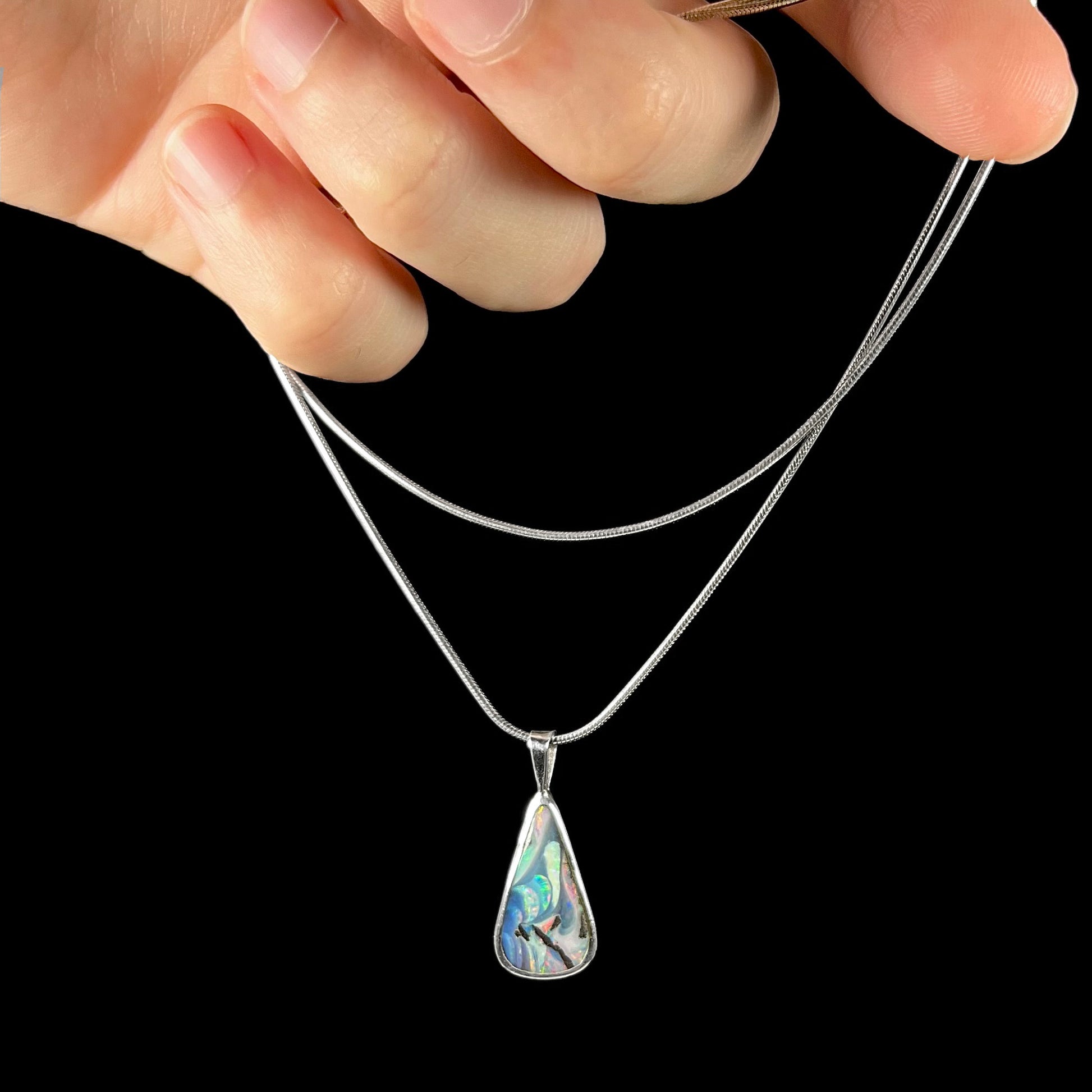 A boulder opal necklace being dangled by the chain in a hand against a black background.