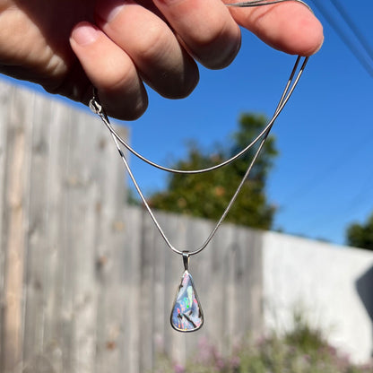 A boulder opal necklace being dangled outdoors in natural light, with pinkish red and green flashes visible.