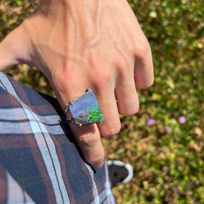 A person resting their hand on their hip in sunlight while wearing a boulder opal ring with bright green flashes.