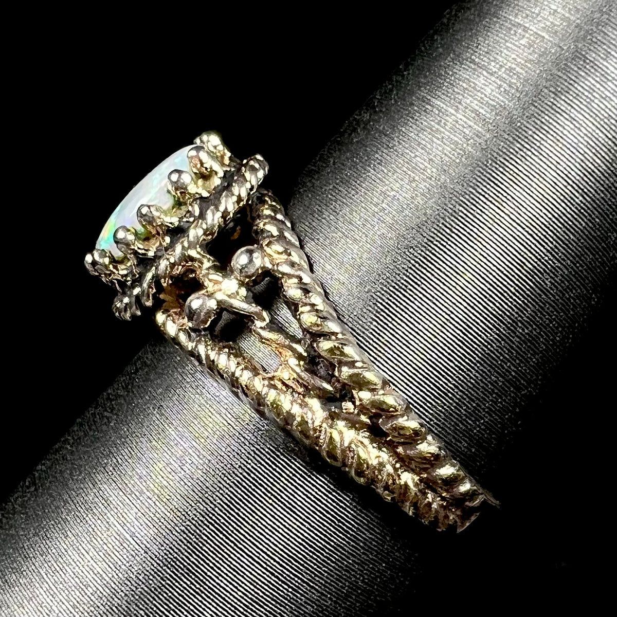 A brass Australian opal ring, shown from the side view on a ring display; the ring's twisted rope-design shank is in focus, as are the regal-style prongs that hold the opal.