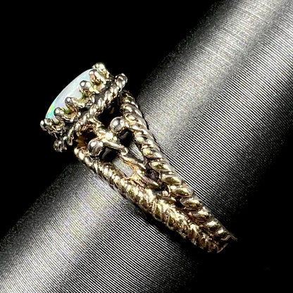 A brass Australian opal ring, shown from the side view on a ring display; the ring's twisted rope-design shank is in focus, as are the regal-style prongs that hold the opal.