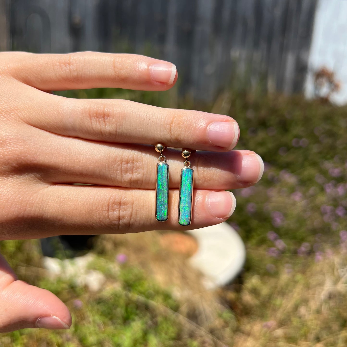 A pair of bright blue opal drop earrings made from yellow gold, shown on top of a hand at a distance in sunlight.