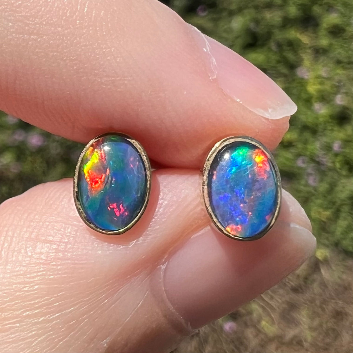 Close-up macro of bright Australian opal triplet stud earrings in 9k gold with bright red and blue flashes of color, held between fingers in natural light
