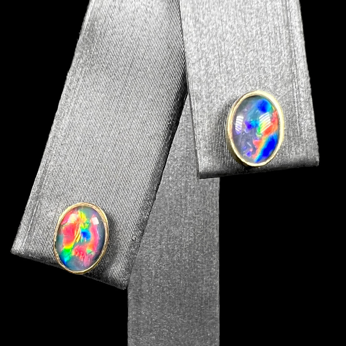 Pair of oval shaped opal triplet stud earrings in 9k gold with bright red and blue flashes of color, shown on earring display