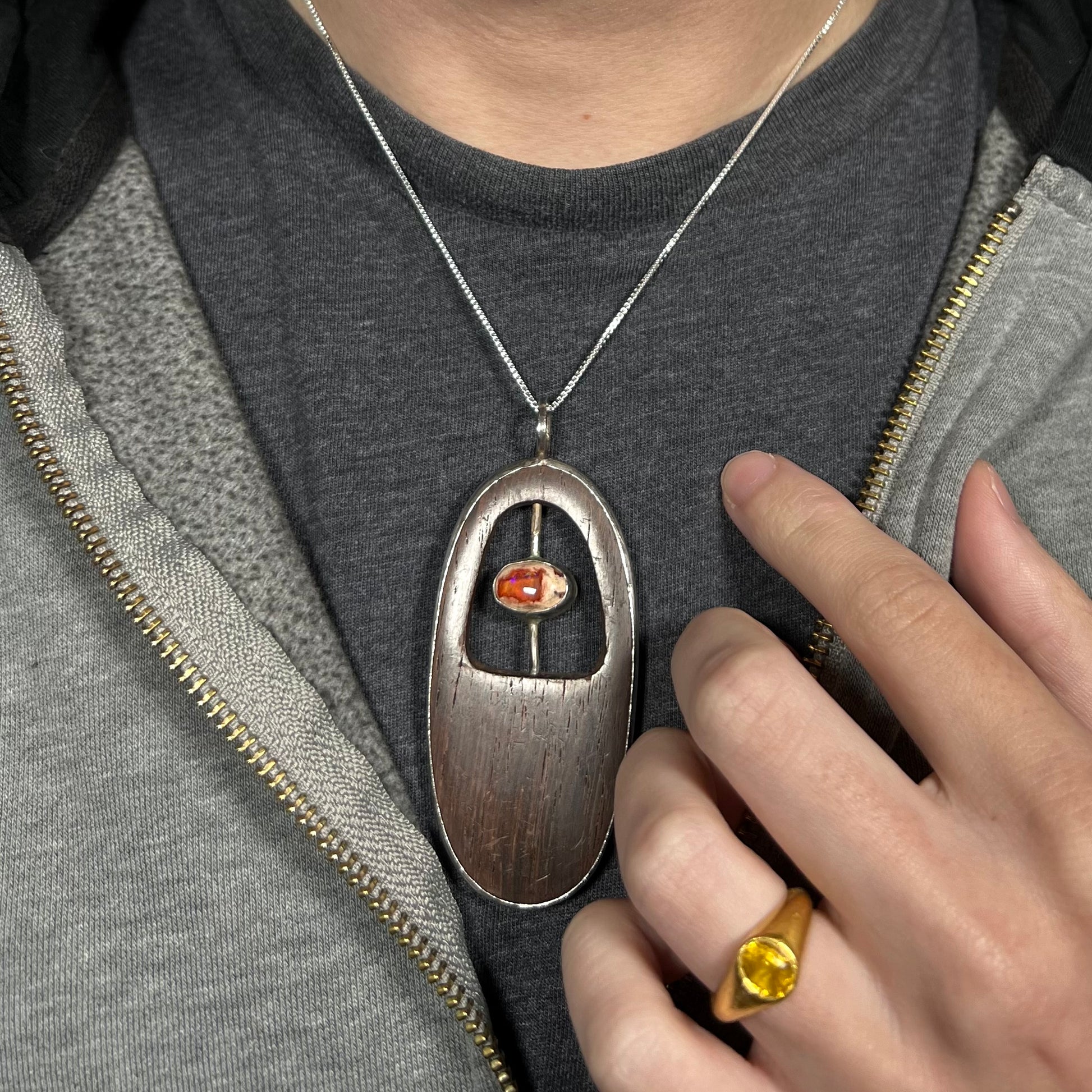 Close-up of a brown wood and orange Mexican fire opal men's necklace in sterling silver, worn around a person's neck against dark shirt and under hoodie.