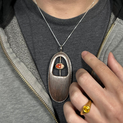 Close-up of a brown wood and orange Mexican fire opal men's necklace in sterling silver, worn around a person's neck against dark shirt and under hoodie.
